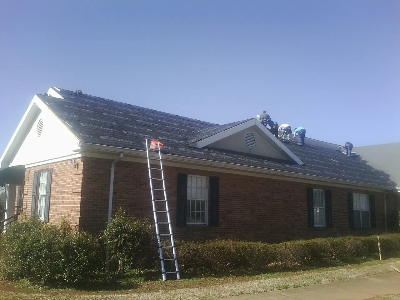 Crew tearing shingles off a brick ranch roof with a ladder set — Emergency Roof Repair work by our crew in Rolling Prairie