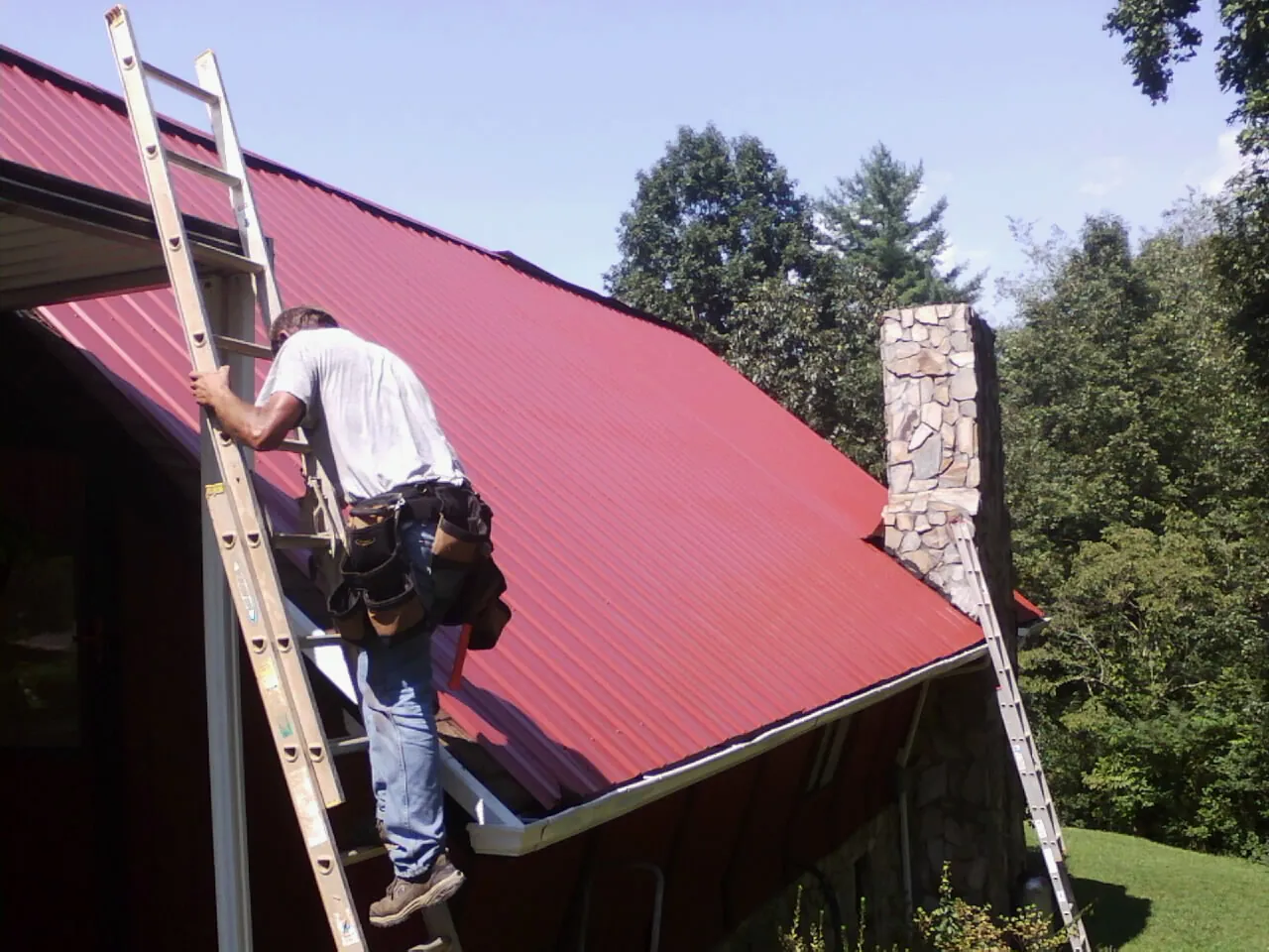 Roofer on a ladder beside a red metal roof and stone chimney — Soffit & Fascia Repair by Roofing Brothers in Rolling Prairie